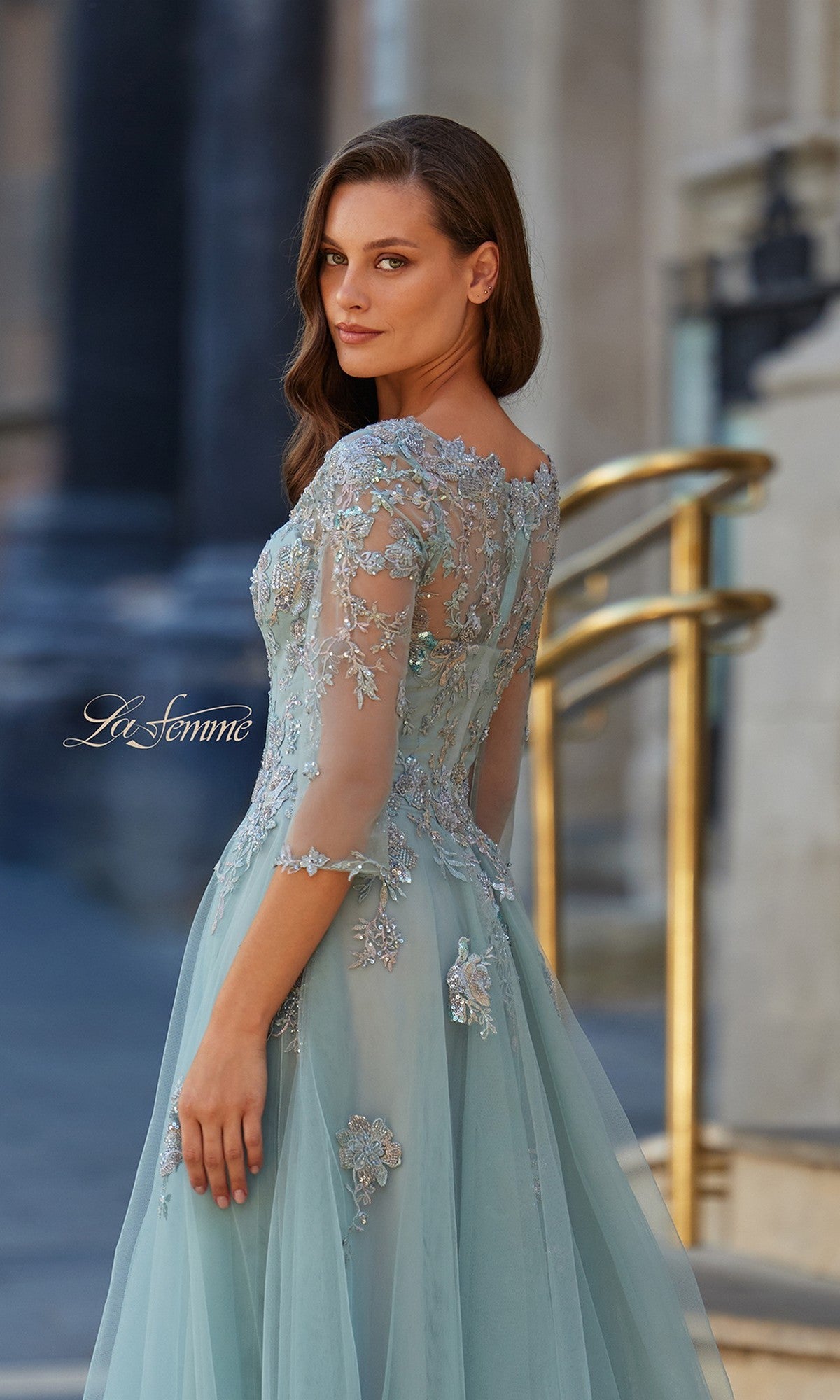 A woman wears the La Femme 33536 long formal dress, featuring light blue tulle, sheer sleeves, and floral embroidery, while standing outdoors near golden railings and stone buildings.